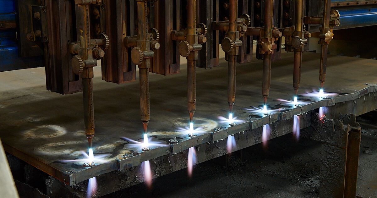 Close-up of multiple flame cutting torches in operation, emitting bright blue flames as they cut through thick metal plates.